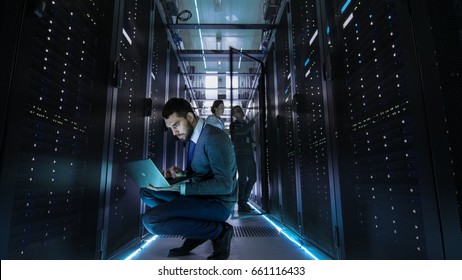 IT Technician Works On Laptop In Big Data Center Full Of Rack Servers. Multiple People Works At Data Center At The Same Time.