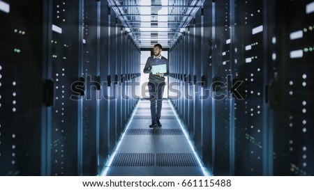 IT Technician Works on Laptop in Big Data Center full of Rack Servers. He Runs Diagnostics and Maintenance, Sets System Up.