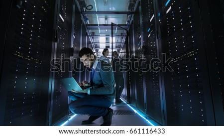IT Technician Works on Laptop in Big Data Center full of Rack Servers. Multiple People Works at Data Center at the same time.