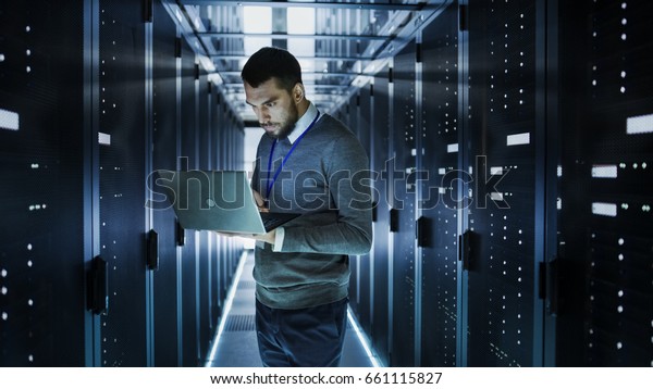 IT Technician Works on a Laptop in Big Data\
Center full of Rack Servers. He Runs Diagnostics and Maintenance,\
Sets up System.