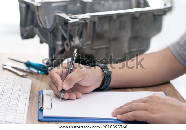 Technician writing on the white paper\
clip board file and check damaged point of the old oil pan parts\
check and repair damaged engines and service\
concept