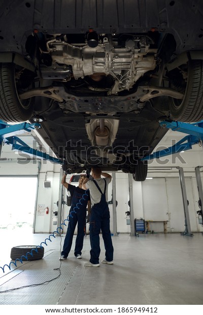 Technicians in car service station uniform\
working together with machine bottom using flashlight and\
screwdriver, vertical shot. Car repairing\
concept