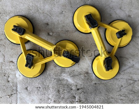A technician's tool with glass expertise called a glass sucker to lift. 3 legs glass sucker painted in yellow. A top-down look of two yellow glass suckers placed on an old foam floor.
