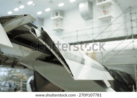 Technicians are working inside a spacious hangar on an aircraft's wing. The area is brightly lit and filled with scaffolding. Repair operations are underway to ensure safety.
