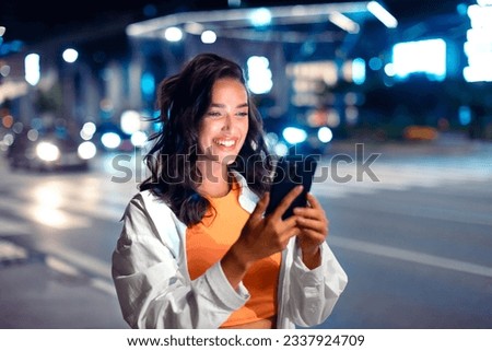 Techno night walks. Smiling woman holding smartphone, reading message while walking through night city street full of neon lights, free space. Excited lady with cellphone outdoors
