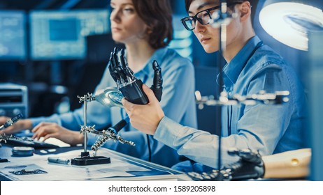 Technological Prosthetic Robot Arm Is Tested By Two Professional Development Engineers In A High Tech Research Laboratory With Modern Futuristic Equipment. Man And Female Compare Data On A PC