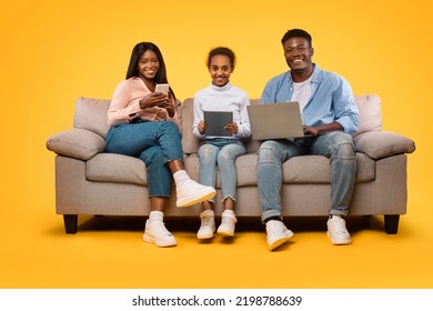 Technologies For Family. Happy Black Mom, Dad And Their Daughter Sitting On Couch With Different Gadgets, Yellow Studio Background. Parents And Child Using Computer, Digital Tablet And Cellphone