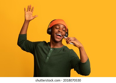 Technologies For Fun. Cheerful black hipster guy listening music in wireless headphones and singing, positive young african man using smartphone as microphone, having fun on yellow background - Powered by Shutterstock