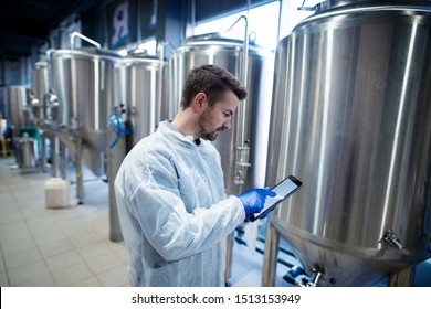 Technologist Expert Standing In Food Production Plant And Typing On His Tablet Computer. Industrial Worker Controlling Production And Checking Quality.