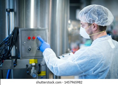 Technologist In White Protective Uniform With Hairnet And Mask Operating On Industrial Machine For Food Processing. Industrial Worker Pushing Button On Machine And Controlling Production.