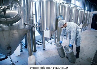 Technologist Working In Factory Handling Chemicals In Production Plant. Worker In Protective Suit Carrying Chemicals In Metal Reservoirs.