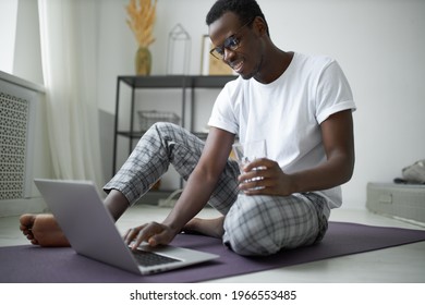 Technology, activity and daily routine concept. Attractive young dark skinned freelancer sitting on fitness mat in front of open laptop, checking email, having glass of water after morning training