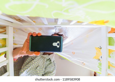 Technology Addition And Parenthood Concept. Perspective Of Lying Baby In Baby Cot. Young Dad Is  Taking Photo With Cellphone.