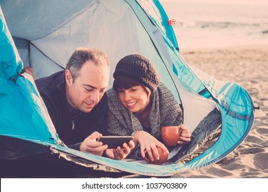 Technology Alternative Vacation Concept For A Middle Aged Couple Looking The Smart Phone Inside A Tent On The Beach In Tenerife. Ocean In Background And Warm Filter With Blue Colors.