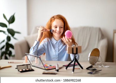 Technology And Beauty Blog For Youth. Portrait of smiling vlogger girl doing makeup, applying and putting face powder with brush, looking in the mirror, recording video using smartphone on tripod