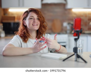 Technology And Blogging Concept. Redhair Woman Recording Video Concent For Her Blog At Home And Looking At Smartphone On Tripod At Kitchen Table, Streaming Online Podcast On Mobile Phone.