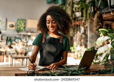 Technology for business. Beautiful african woman using wireless laptop and writing on clipboard at modern flower boutique. Positive female owner analyzing stock of plants at favorite small business. - Powered by Shutterstock