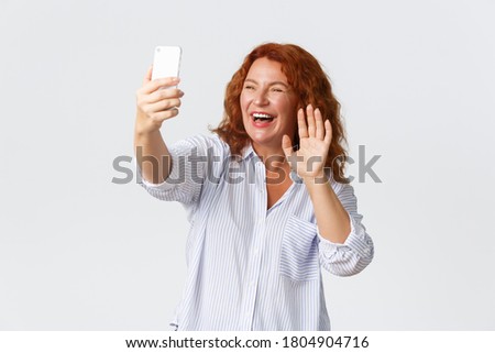 Technology and communcation concept. Friendly and outgoing smiling, attractive middle-aged redhead woman talking on videochat, holding cellphone, waving hello at mobile phone and laughing