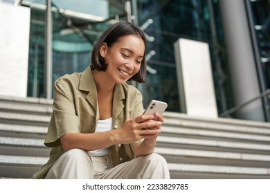Technology and communication. Young smiling girl, asian woman sits with smartphone, reads message with big smile. - Shutterstock ID 2233829785