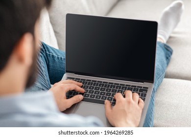 Technology concept. Unrecognizable guy using laptop computer with blank screen, surfing net and advertising internet website, sitting at home on sofa, over the shoulder view. Mockup, selective focus