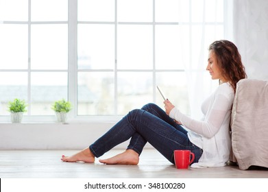 Technology and coziness. Beautiful young woman with cup of tea using tablet computer while sitting on floor at home. - Powered by Shutterstock
