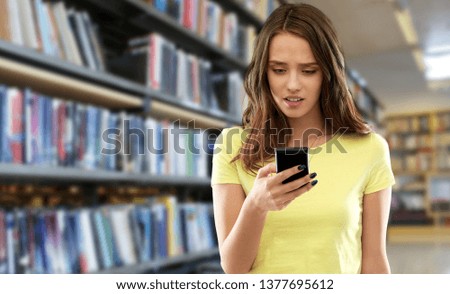 technology, cyberbullying and people concept - young woman or teenage girl in blank yellow t-shirt using smartphone over library background