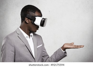 Technology, entertainment, gaming and cyberspace. African office worker in formal suit experiencing virtual reality using 3d headset for mobile phone, looking at his open palm as if holding something - Powered by Shutterstock