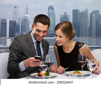 Technology, Food, Holidays And People Concept - Smiling Couple With Smartphone Eating At Restaurant Over City Background