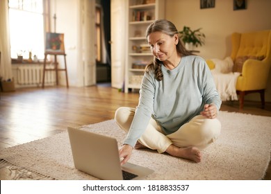 Technology, Gadgets And Aging Concept. Barefoot European Female Pensioner Using Portable Computer At Home, Siting On Carpet, Surfing Internet Or Watching Online Yoga Video Tutorial For Aged People