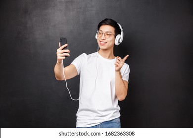 Technology, Gadgets And People Concept. Portrait Of Stylish Young Asian Hipster Guy In Headphones, Video-call Friend Using Smartphone, Taking Selfie Pointing Finger Right, Black Background