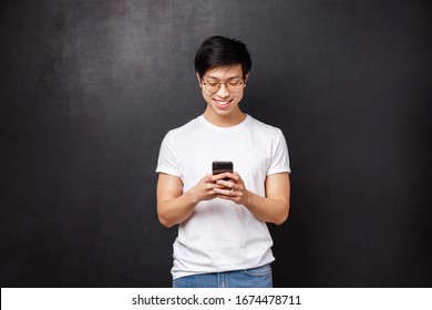 Technology, Gadgets And People Concept. Smiling Teenage Asian Guy Playing On Phone, Texting Friend Using Smartphone Messanger, Use New App To Edit Photos Social Media, Black Background