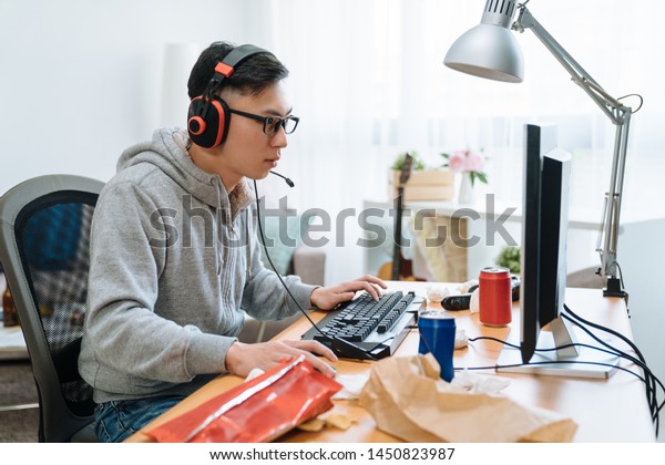 technology gaming entertainment concept. happy young\
asian man in headphones with pc computer playing game and streaming\
playthrough video. lazy college boy relax home on summer break eat\
junk food