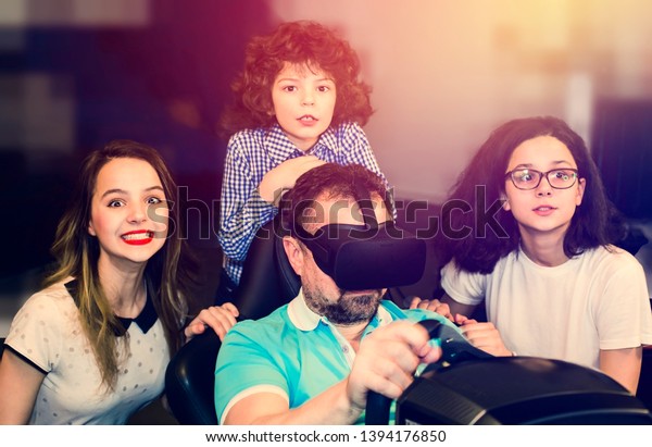 technology,\
gaming, entertainment and people concept. Happy young man is\
playing racing videogame in 3D virtual reality simulator using\
headset. Happy family having fun\
together.