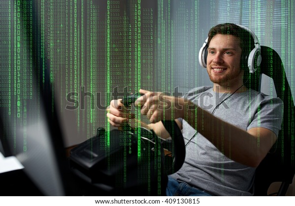 technology, gaming, entertainment\
and people concept - happy smiling young man in headphones with pc\
computer playing car racing video game at home and steering\
wheel