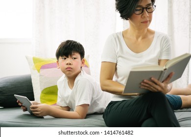 Technology And Generation Gap. Book And EBook. Smart Looking Preteen Son Reading Ebook In Tablet, Peaking At Good Looking Asian Mother's Book With Curiosity About Book His Mom Is Reading. 