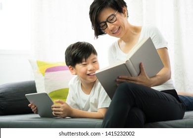 Technology And Generation Gap. A Good Looking Asian Mother And Son Sharing A Beautiful Moment Of Reading Together In Living Room Full Of Joy And Happiness. Books VS EBook, Contrast, Diversity Concept.