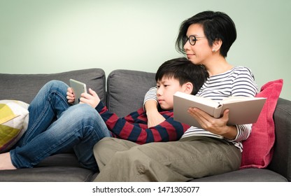 Technology And Generation Gap. Good Looking Asian Mother Sit Relaxing With Her Smart Preteen Son's, Sharing A Beautiful Moment Of Reading Together On A Couch In Living Room. Books VS EBook Concept.