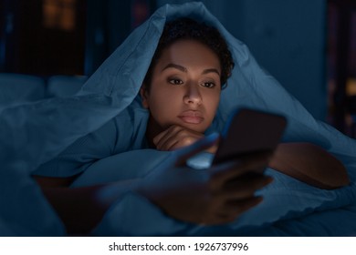 technology, internet, communication and people concept - young african american woman with smartphone lying under blanket in bed at home at night - Shutterstock ID 1926737996