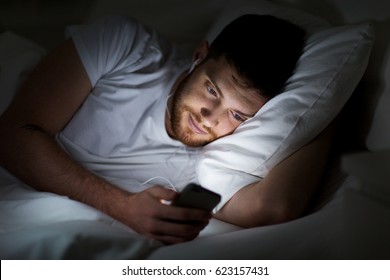 technology, internet, communication and people concept - happy smiling young man with smartphone and earphones listening to music in bed at night - Powered by Shutterstock