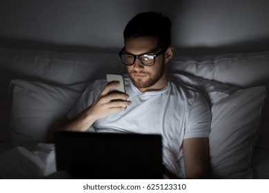 technology, internet, communication and people concept - young man in glasses with laptop computer and smartphone in bed at home bedroom at night