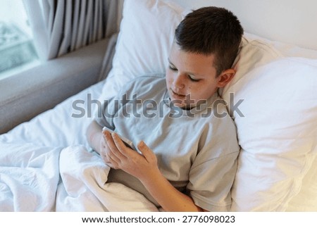  Technology and internet concept. Boy laying on bed and looking at smartphone. Child playing with mobile phone, lying on a bed. Freetime.