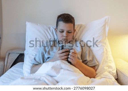  Technology and internet concept. Boy laying on bed and looking at smartphone. Child playing with mobile phone, lying on a bed. Freetime.