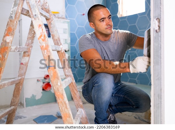 The technology of laying tile, repair and\
decoration. Focused man wearing ordinary clothes puts porcelain\
tiles on wall sitting squat at\
bathroom