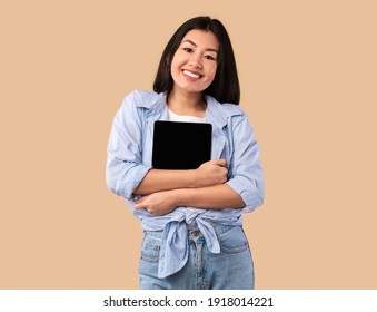 Technology Lover Concept. Happy Asian Woman Hugging Tablet With Black Blank Empty Screen, Holding It Tight Near Chest. Casual Female Geek Posing Isolated Over Studio Background Wall, Copy Space