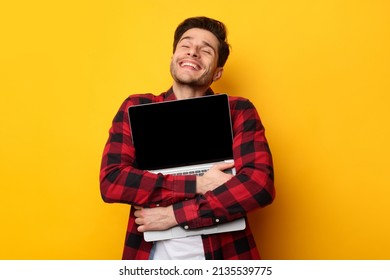 Technology Lover Concept. Happy man hugging laptop with black blank empty screen, holding it tight near chest. Casual male geek with closed eyes standing isolated on yellow orange studio wall