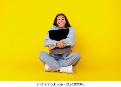 Technology Lover Concept. Happy Woman Hugging Laptop With Black Blank Empty Screen, Holding It Tight Near Chest. Casual Female Geek Sitting On Floor With Closed Eyes Isolated On Yellow Studio Wall