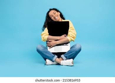 Technology Lover Concept. Happy Woman Hugging Laptop With Black Blank Empty Screen, Holding It Tight Near Chest. Casual Female Geek Sitting On Floor With Closed Eyes Isolated On Blue Studio Wall