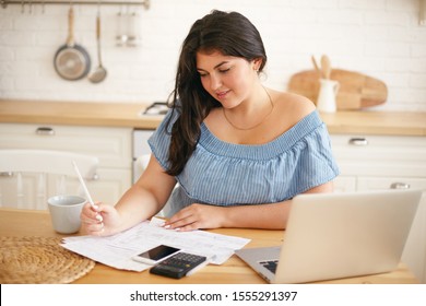 Technology, occupation and education. Beautiful college girl doing home assignment in kitchen, using laptop. Cute chubby woman managing finances at home, sitting at table with electronic devices - Powered by Shutterstock