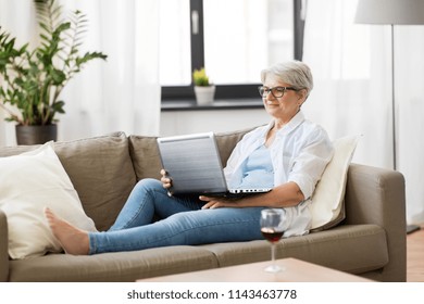 Technology, Old Age And People Concept - Happy Senior Woman In Glasses With Laptop Computer And Glass Of Wine At Home