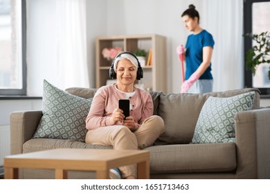 technology, old age and people concept - happy senior woman in headphones with smartphone and housekeeper cleaning at home
